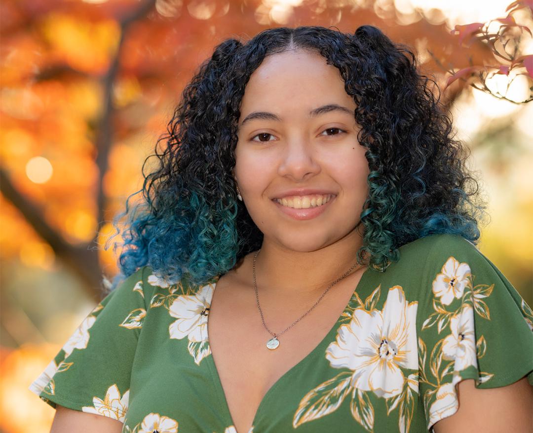 Smiling headshot of a woman with brightly colored, curly hair with trees behind her