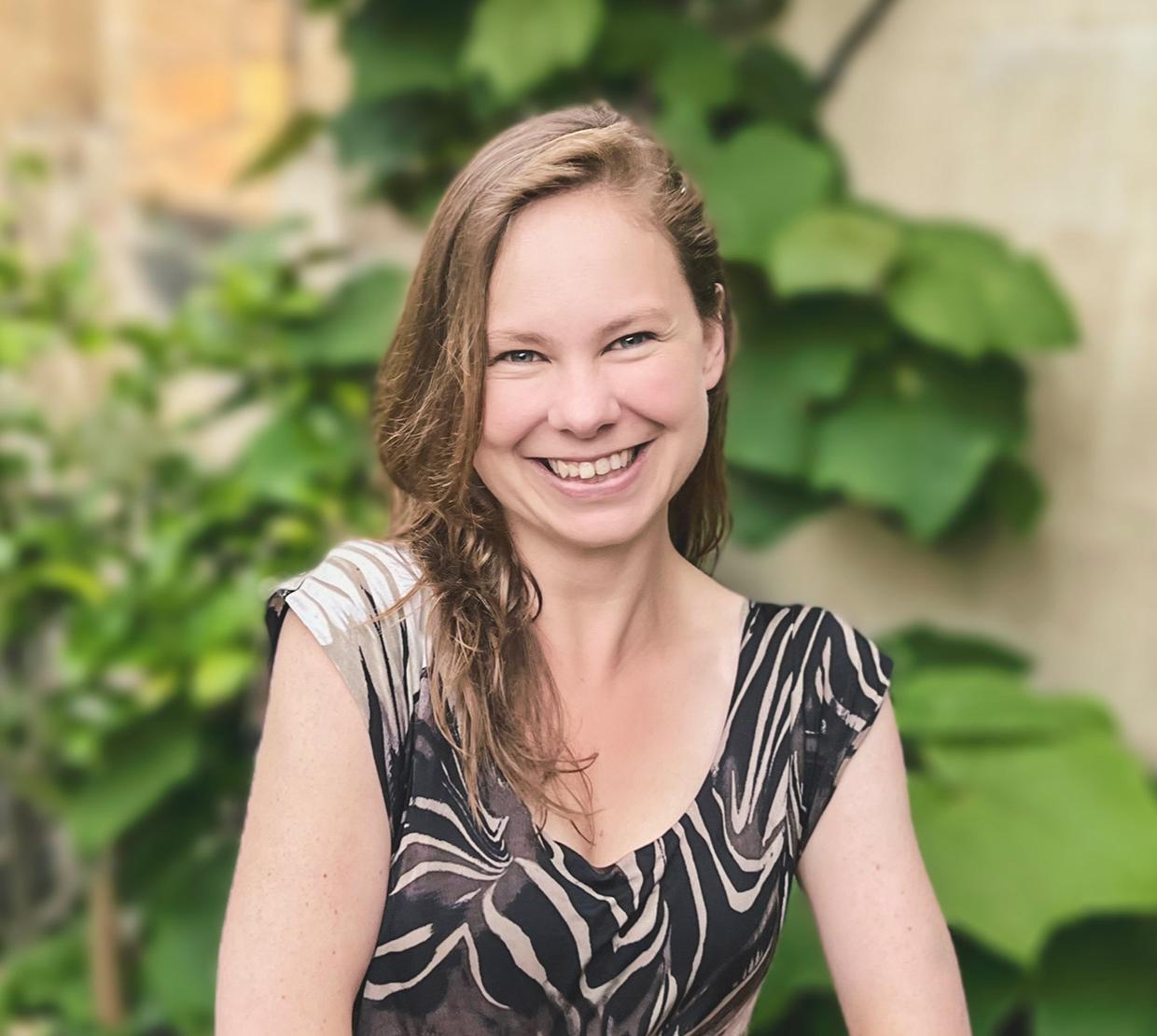 Headshot of Dr. Laura P. Schaposnik: A woman smiling with her hands folded in front of her, sitting outside a house of stone walls and healthy plants.