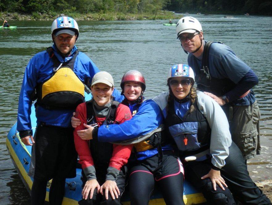 Mary Tunstall smiles with her rafting team. She sits in the middle wearing a blue jacket and hugging a friend.