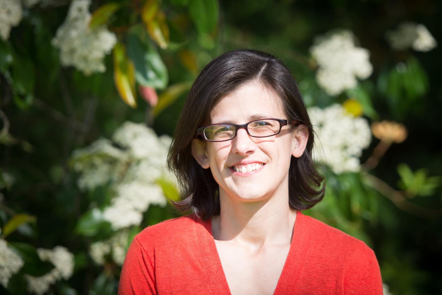 A woman in a red shirt with dark hair stands in front of a green leafy background with white flowers.