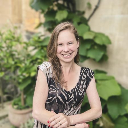 Headshot of Dr. Laura P. Schaposknik: A woman smiling with her hands folded in front of her, sitting in front of a background featuring beige stone walls and green plants.