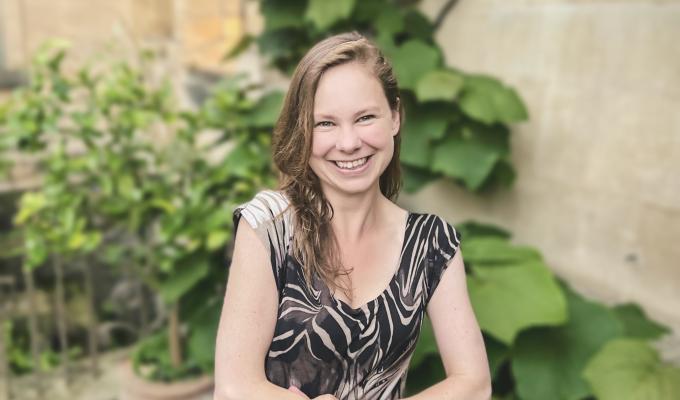 Headshot of Dr. Laura P. Schaposknik: A woman smiling with her hands folded in front of her, sitting in front of a background featuring beige stone walls and green plants.