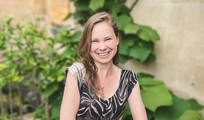 Headshot of Dr. Laura P. Schaposknik: A woman smiling with her hands folded in front of her, sitting outside a house of stone walls and healthy plants.