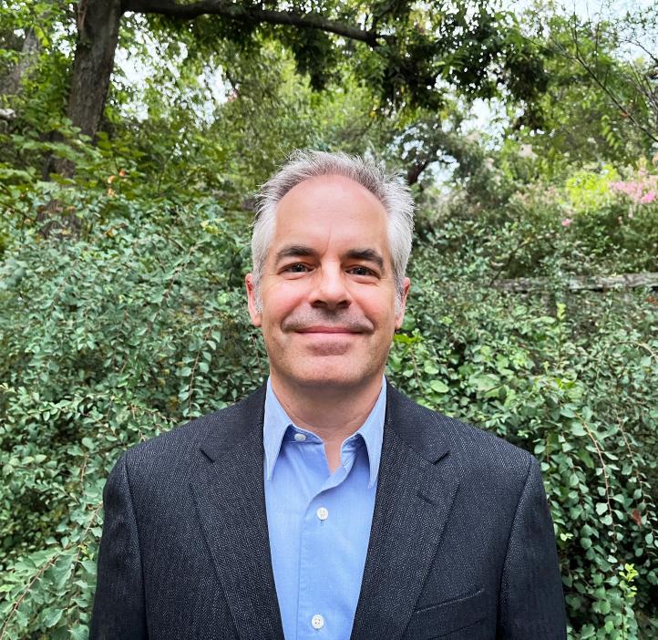 Professor Jon Kujawa stands in a suit coat and smiles in his headshot outdoors surrounded by greenery. 