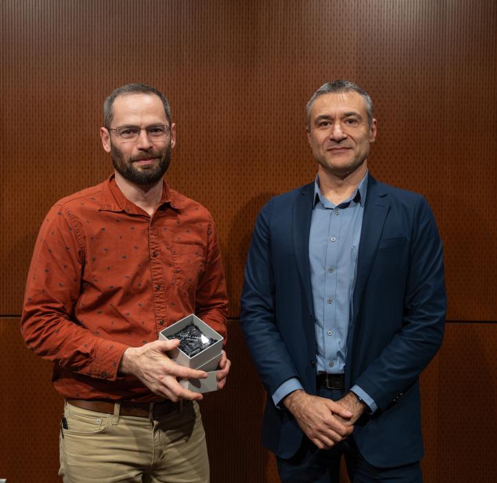 A man in an orange shirt holds a glass award standing next to a man in a blue suit.