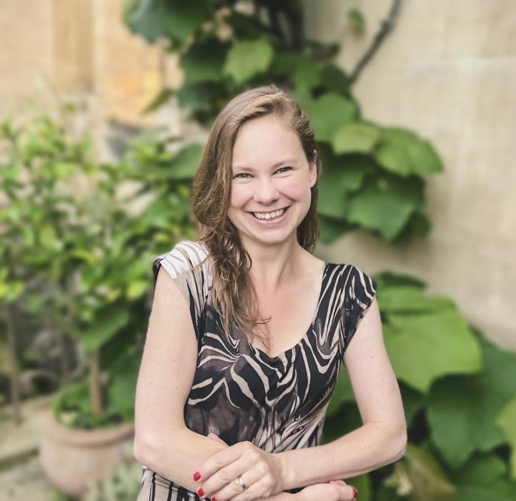 Headshot of Dr. Laura P. Schaposknik: A woman smiling with her hands folded in front of her, sitting in front of a background featuring beige stone walls and green plants.