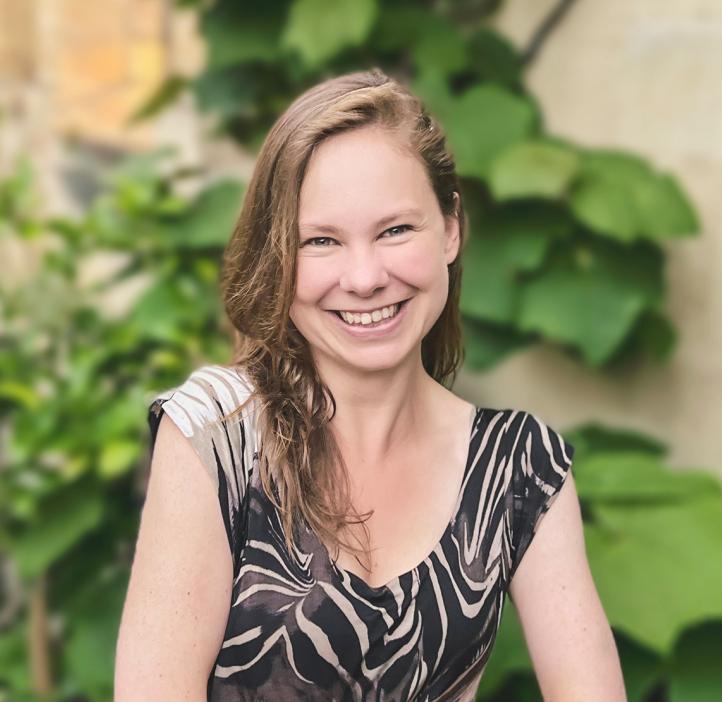 Headshot of Dr. Laura P. Schaposnik: A woman smiling with her hands folded in front of her, sitting outside a house of stone walls and healthy plants.